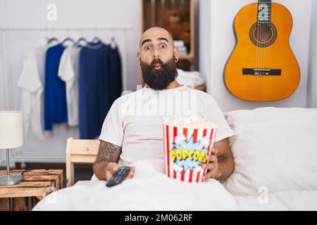 Young hispanic man with beard and tattoos eating popcorn in the bed making fish face with mouth and squinting eyes, crazy and comical. Stock Photo
