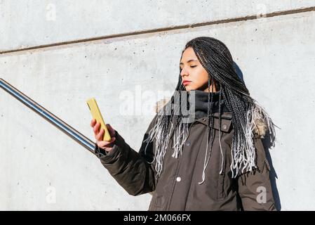 Peaceful female with braided hair browsing social media on cellphone while standing near wall in street on sunny day Stock Photo