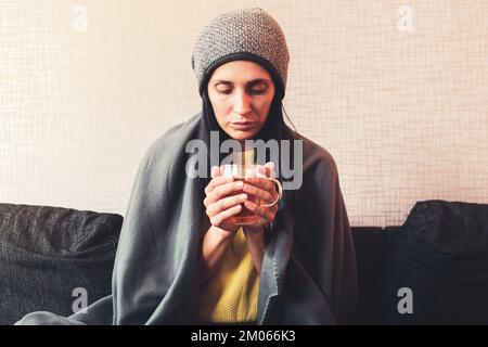 Caucasian Worried sad woman sits under blanket on sofa with cup of tea, Rising costs in private households for gas bill due to inflation and war, Ener Stock Photo