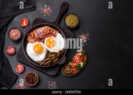 Tasty breakfast consists of eggs, bacon, beans, tomatoes, with spices and herbs on a dark concrete background Stock Photo