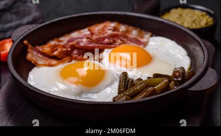 Tasty breakfast consists of eggs, bacon, beans, tomatoes, with spices and herbs on a dark concrete background Stock Photo