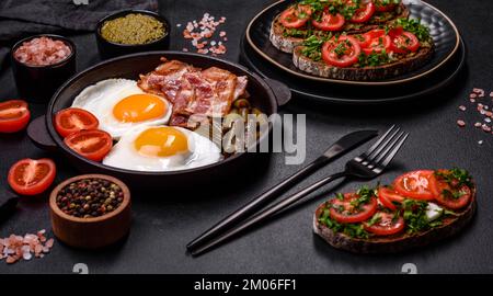 Tasty breakfast consists of eggs, bacon, beans, tomatoes, with spices and herbs on a dark concrete background Stock Photo