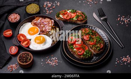 Tasty breakfast consists of eggs, bacon, beans, tomatoes, with spices and herbs on a dark concrete background Stock Photo