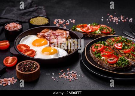 Tasty breakfast consists of eggs, bacon, beans, tomatoes, with spices and herbs on a dark concrete background Stock Photo