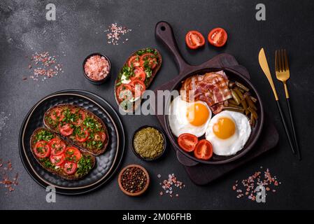 Tasty breakfast consists of eggs, bacon, beans, tomatoes, with spices and herbs on a dark concrete background Stock Photo