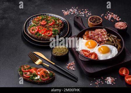 Tasty breakfast consists of eggs, bacon, beans, tomatoes, with spices and herbs on a dark concrete background Stock Photo