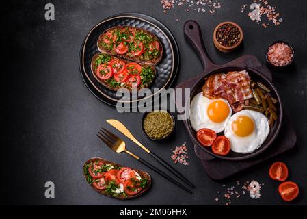 Tasty breakfast consists of eggs, bacon, beans, tomatoes, with spices and herbs on a dark concrete background Stock Photo