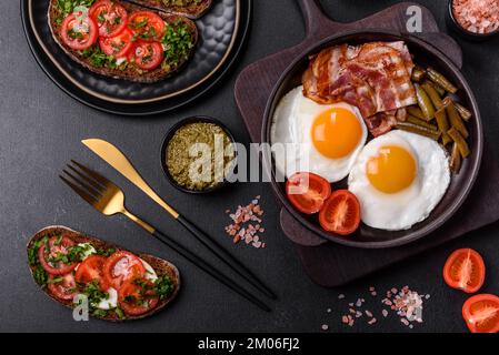 Tasty breakfast consists of eggs, bacon, beans, tomatoes, with spices and herbs on a dark concrete background Stock Photo