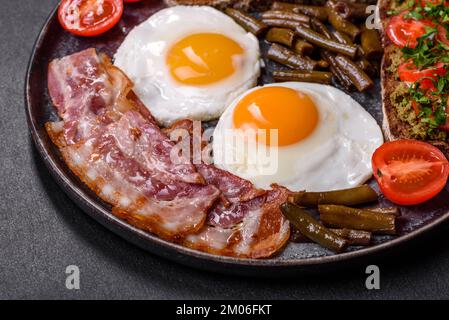 Tasty breakfast consists of eggs, bacon, beans, tomatoes, with spices and herbs on a dark concrete background Stock Photo