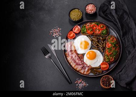 Tasty breakfast consists of eggs, bacon, beans, tomatoes, with spices and herbs on a dark concrete background Stock Photo