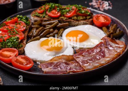 Tasty breakfast consists of eggs, bacon, beans, tomatoes, with spices and herbs on a dark concrete background Stock Photo