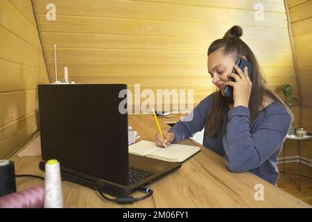 Woman fashion designer working on a laptop, making notes in a notebook, talking on the phone with a client. Small business. Stock Photo
