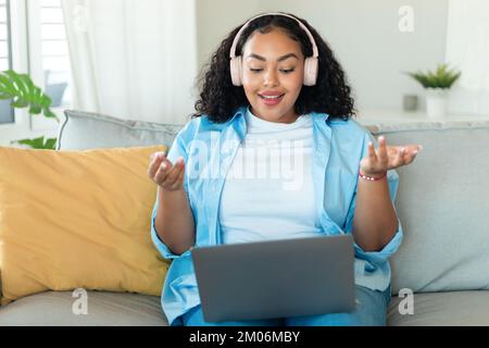 Video conference. Portrait of positive black chubby woman in wireless headset having web call via laptop sitting on sofa Stock Photo