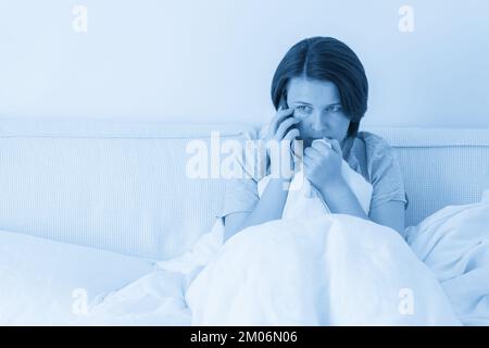 Woman using smartphone as she sits in bed covered with duvet, scared and terrified Stock Photo