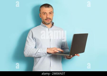 Confident middle aged man holding and using laptop computer, standing over blue studio background Stock Photo