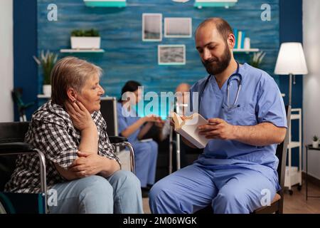 Social worker in a nursing home reading a book for a woman patient in a wheelchair. Elderly female retirement center resident listening carefully to male medical assistant. Stock Photo