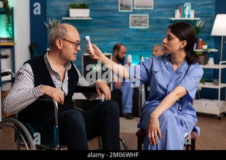 Nurse in a specialized health center measuring the temperature of an elderly patient in a wheelchair, using a digital infrared thermometer. Retired man attending medical consultation with specialists. Stock Photo