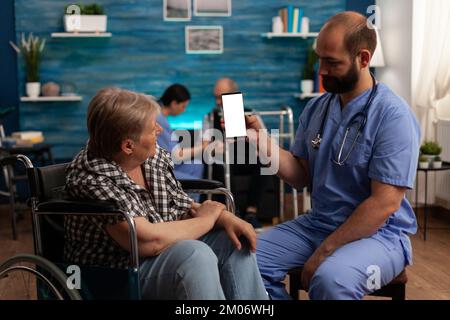 Elderly woman seated in wheelchair next to medical assistant looking at blank display on mobile device. Male nurse holding white screen on smart phone in nursing home, professional care. Stock Photo