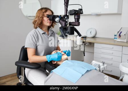 Female dentist with dental tools and microscope treating patient teeth at dental clinic office. Medicine, dentistry and health care concept. Stock Photo