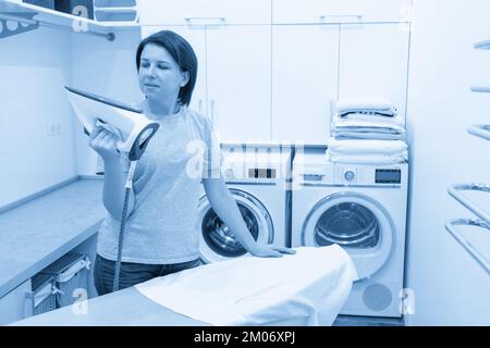 Woman looking at iron in laundry room with washing machine on background Stock Photo