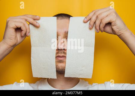 A handsome guy in a protective mask hides behind a wall of toilet paper to prevent coronavirus infection. Yellow background. Flash concept COVID-19. H Stock Photo