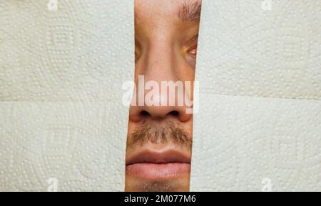 A handsome guy in a protective mask hides behind a wall of toilet paper to prevent coronavirus infection. Yellow background. Flash concept COVID-19. H Stock Photo