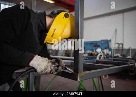 A worker uses a welding machine to weld two metal bars in a workshop and wears a protective visor Stock Photo
