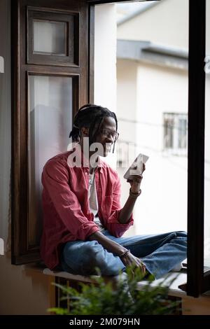 Happy carefree African American man sits on windowsill of apartment chatting with friends via phone Stock Photo