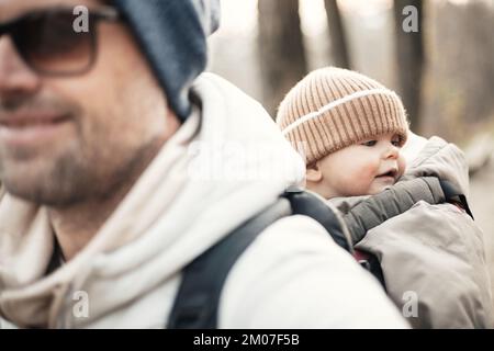 Sporty father carrying his infant son wearing winter jumpsuit and cap in backpack carrier hiking in autumn forest Stock Photo