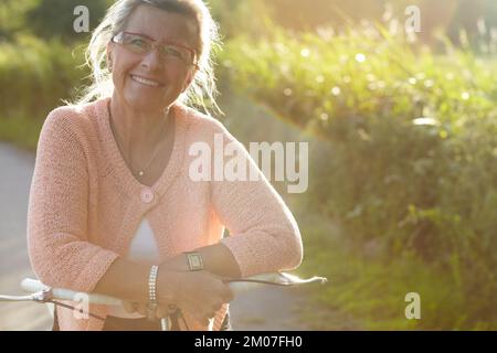 A ride in the evening sunlight. Portrait of a senior woman leaning on her bicycle handles and smiling happily. Stock Photo