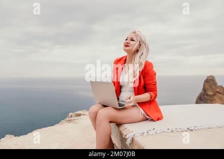 Digital nomad, a business woman in red blazer with a laptop sits on the rocks by the sea during sunset, makes a business transaction online from a Stock Photo