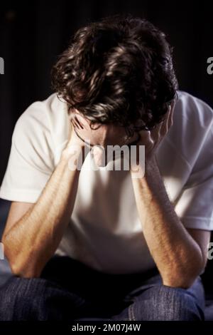 Hes going through a lot. A young man sitting on the foot of his bed with his hands on his face in despair. Stock Photo