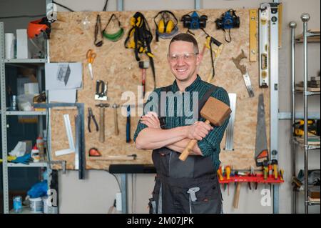 Portrait of a carpenter in goggles and overalls holding a wooden hammer in the workshop against the background of a wall with tools. Stock Photo