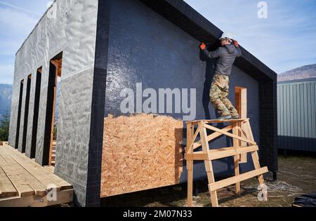 Male painter using paint roller, doing exterior paint work in a black color. Man worker building wooden frame house. Carpentry and construction concept. Stock Photo