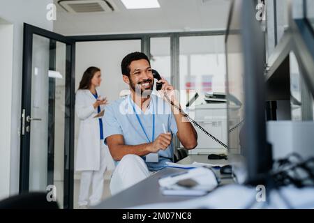 Young multiracial doctor sitting at reception and calling, his colleagues discussing in the background. Stock Photo