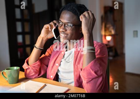 Smiling African guy college student talking on phone while studying at home, taking break from study Stock Photo