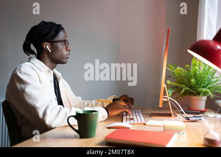 Focused African man freelance web developer writing code, using computer, sitting at home office Stock Photo