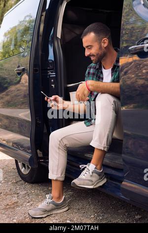 man using his phone sitting in the door of his van Stock Photo