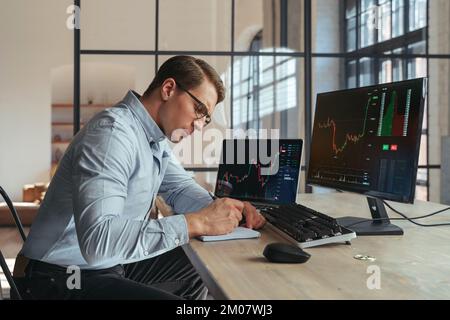 Businessman crypto trader sitting at office table writing in his notepad, noting down info after checking ico and trading market dynamics on his laptop and personal computer with bitcoin on his desk Stock Photo
