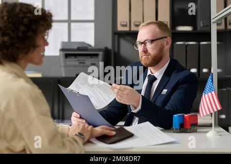 Portrait of bearded male consultant working in US embassy office and holding application form while talking to woman Stock Photo