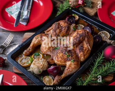 Christmas meat dish with roasted whole chicken with lemon, garlic, onion and herbs. Served on wooden table with red plates, napkins and decoration Stock Photo
