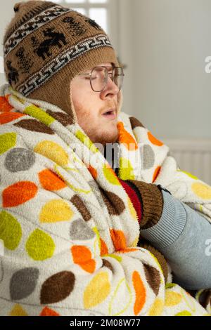 A young man wrapped up in a blanket and wearing a woolly hat indoors Stock Photo