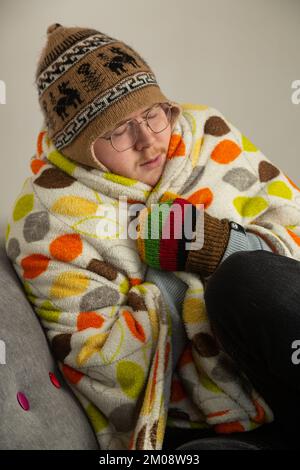 A young man wrapped up in a blanket and wearing a woolly hat indoors Stock Photo