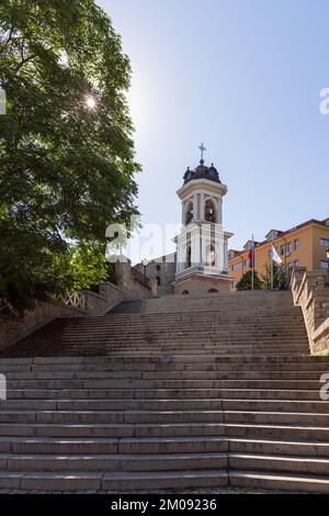 Vertical bottom-up view of three long flights of neat stone stairs, 3-level belltower at top, early morning sun peeks through foliage of tree. Plovdiv Stock Photo