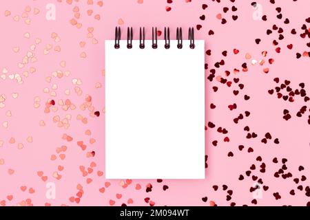 Empty notepad mockup with red metallic hearts confetti scattered on a pink background. Festive compisition. Stock Photo