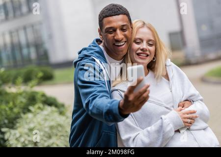 cheerful interracial couple in hoodies taking selfie on mobile phone outdoors Stock Photo