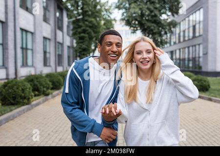 happy multiethnic couple in hoodies holding hands and looking at camera on urban street Stock Photo