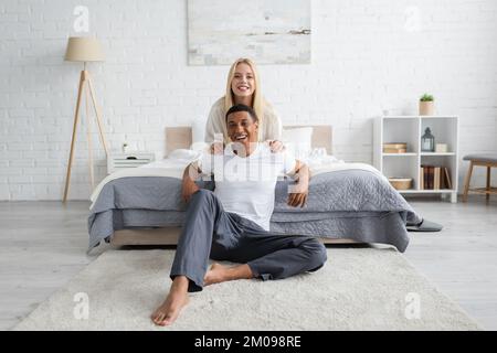 cheerful african american man in sleepwear sitting on floor in bedroom near blonde woman hugging his shoulders Stock Photo