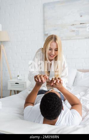 excited blonde woman holding hands of african american boyfriend while having fun in bedroom Stock Photo