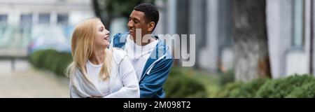 joyful multiracial couple in love looking at each other on urban street, banner Stock Photo